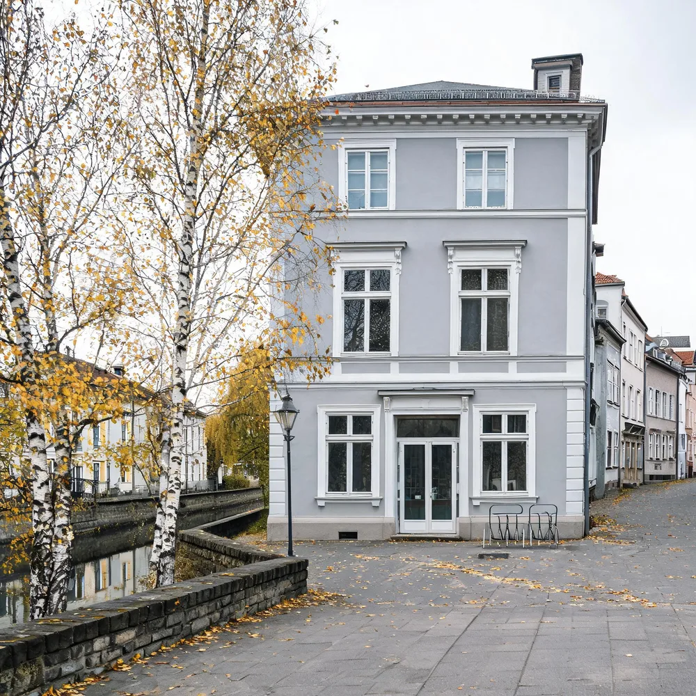 Hellgraues dreistöckiges Stadthaus mit ebenerdiger Glastür und Fahrradbügeln, links Birken und der Fluss hinter einer niedrigen Steinmauer.