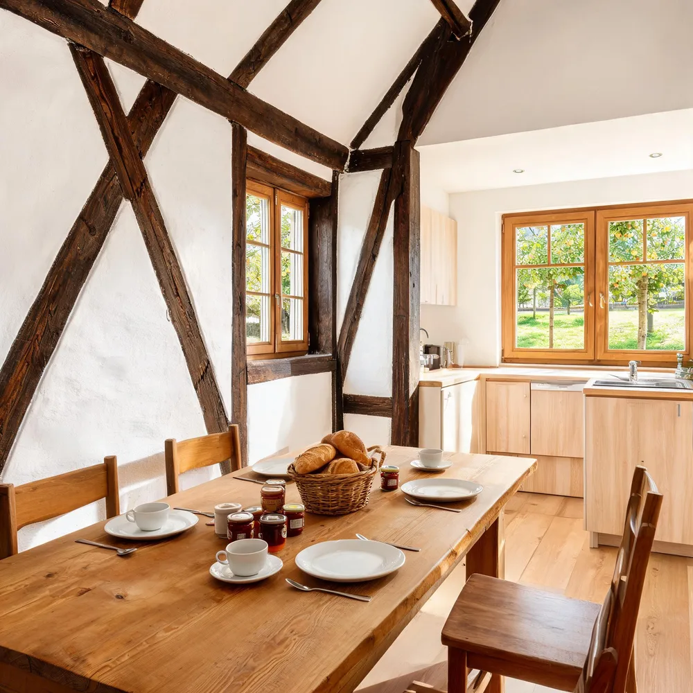 Wohnküche im Fachwerk-Obergeschoss: dunkle Eichenbalken in weißen Wänden, langer Holztisch mit Frühstück für vier, Fenster zum Apfelgarten.
